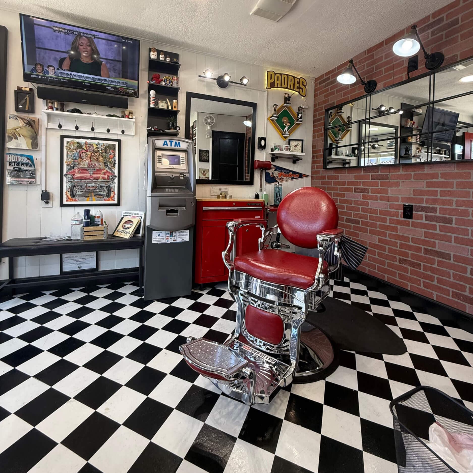 Barbershop interior with a red leather barber chair, black and white checkered floor, TV, ATM, and Padres decor.