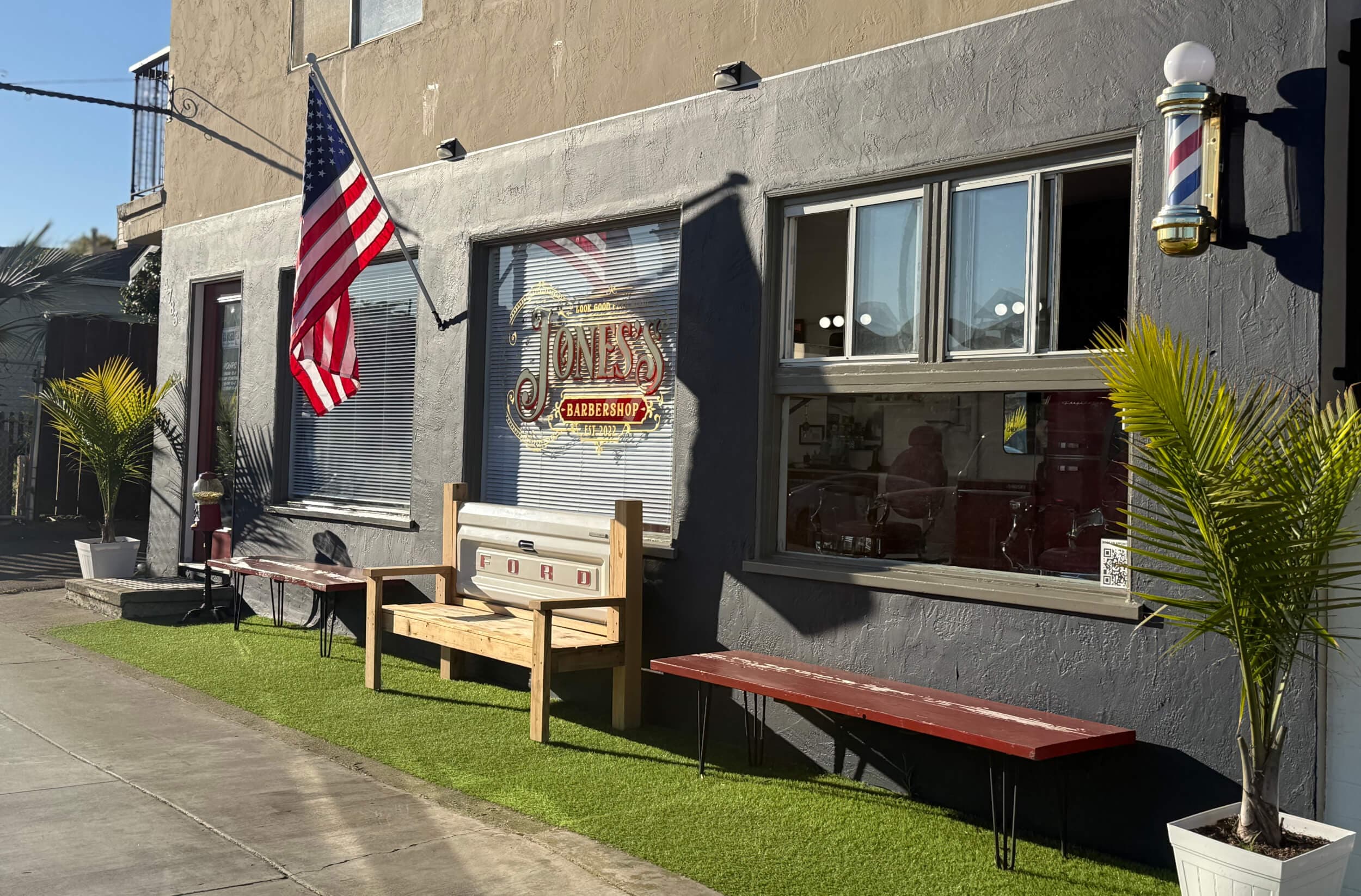 Exterior of Jones's Barbershop with an American flag, barber pole, and benches on artificial turf in sunlight. One bench is made from a Ford tailgate.