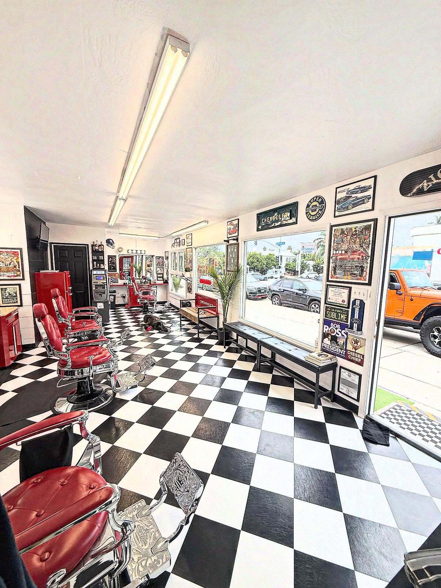 A retro barbershop with a black and white checkered floor, red barber chairs, car-themed decor, and a dog resting.