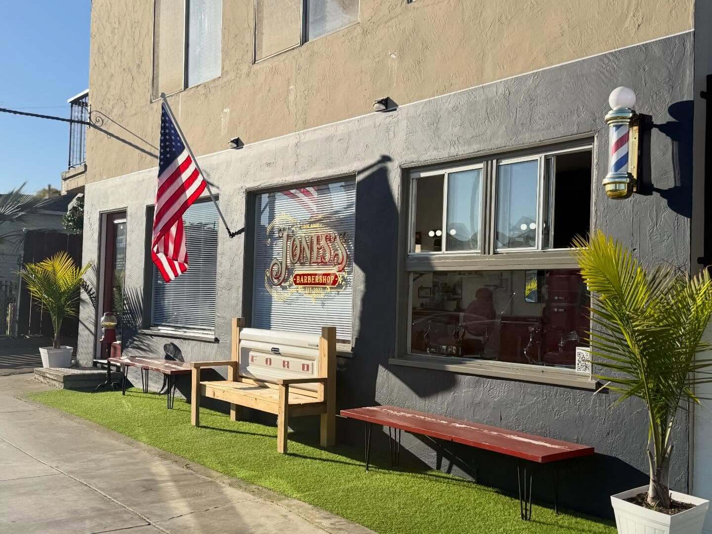 Jones's Barbershop exterior with an American flag, barber pole, artificial turf, and a Ford tailgate bench.