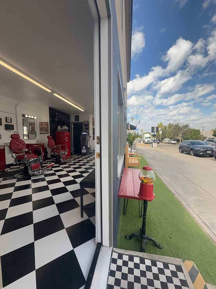 Interior of a barber shop with red chairs and checkerboard floor, looking out to a street with a gumball machine.